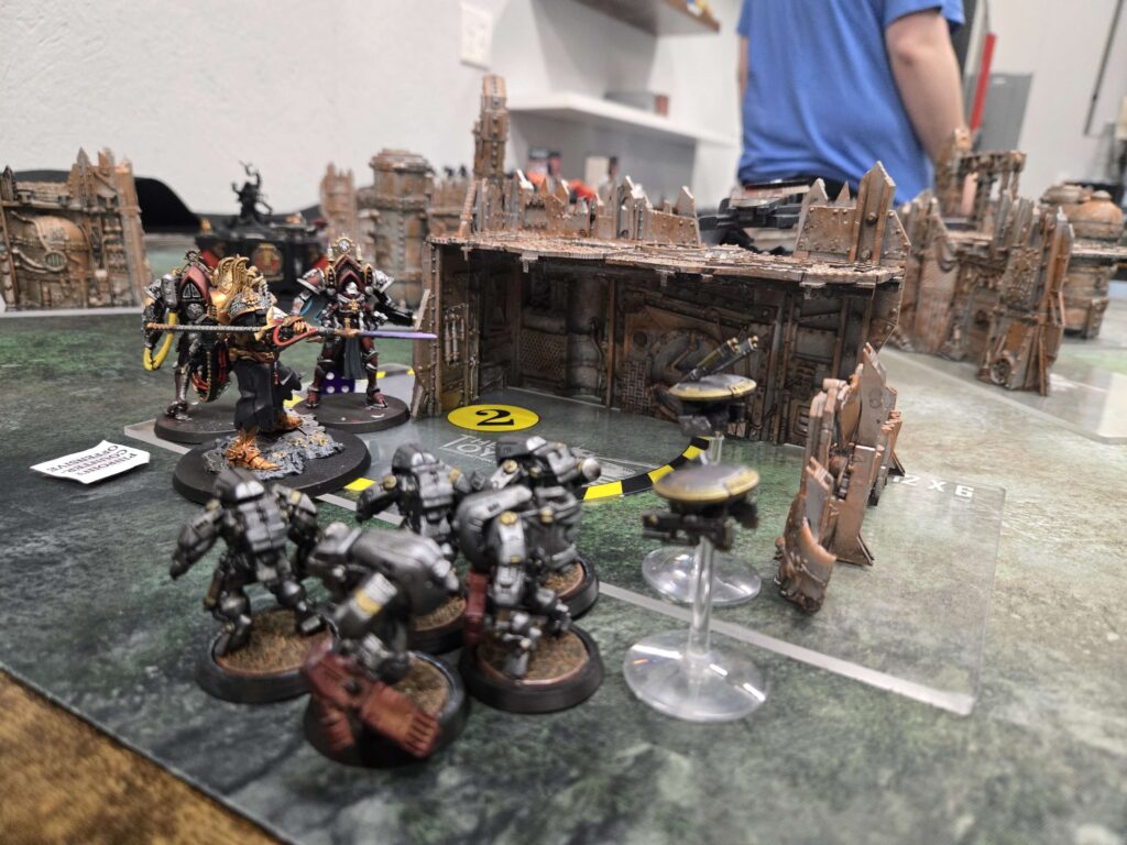 Tabletop wargaming scene with armored miniatures advancing across a ruined city battlefield and rubble-filled buildings in the background.