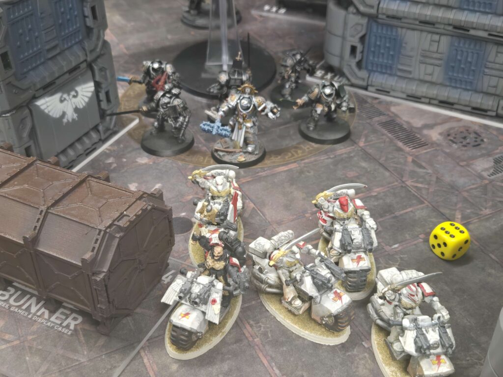 Group of painted Warhammer 40k miniatures in armor posed on a tabletop battlefield with ruins and a yellow dice nearby