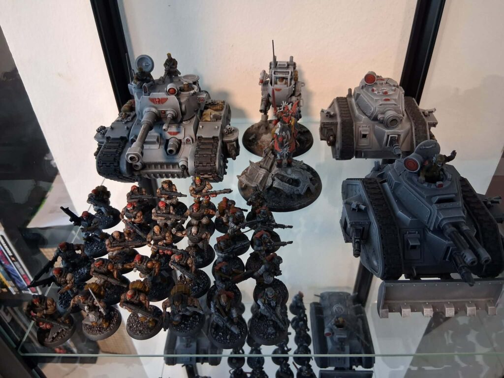 Display shelf with multiple gray armored tanks and infantry miniatures from a tabletop wargame arranged in rows on glass shelves.
