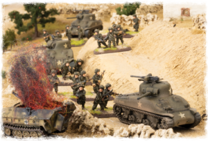 WWII toy diorama: miniature soldiers advance between armored tanks in a sandy, desert-like battlefield with a burning wreck on the left.