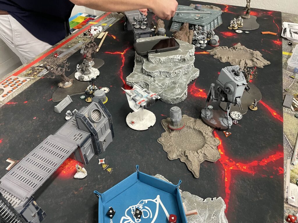 Tabletop wargame in progress with rocky terrain, lava-themed mat, and numerous miniatures; a player moves a model.
