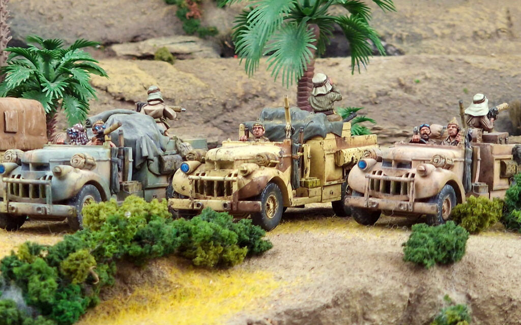 Miniature desert battlefield diorama with dusty toy jeeps and soldiers in helmets hiding among rocks and foliage.