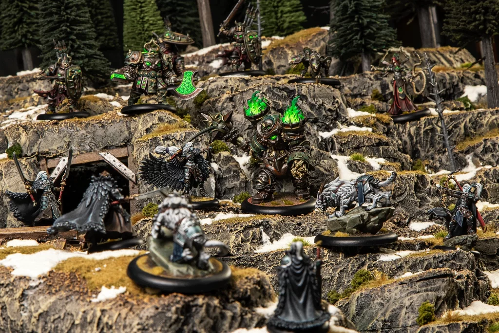 LifestyleACamera67 Frontline Gaming Tabletop fantasy battle scene with armored miniatures on rocky, mossy terrain and glowing green weapons and shields into a forested backdrop.