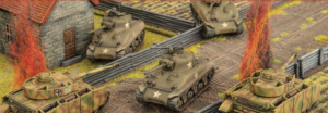 Miniature WWII battle scene with several model tanks in a trench landscape and a small brick building nearbyartillery lines and red brush in the background