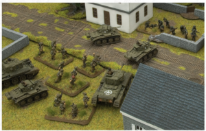 Tabletop wargame diorama with armored vehicles and infantry advancing past a white building on a grassy battlefield.