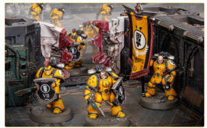 Yellow-armored Space Marines form a battle line on a sci-fi diorama with a black banner featuring a white fist emblem and ruined scenery behind them.