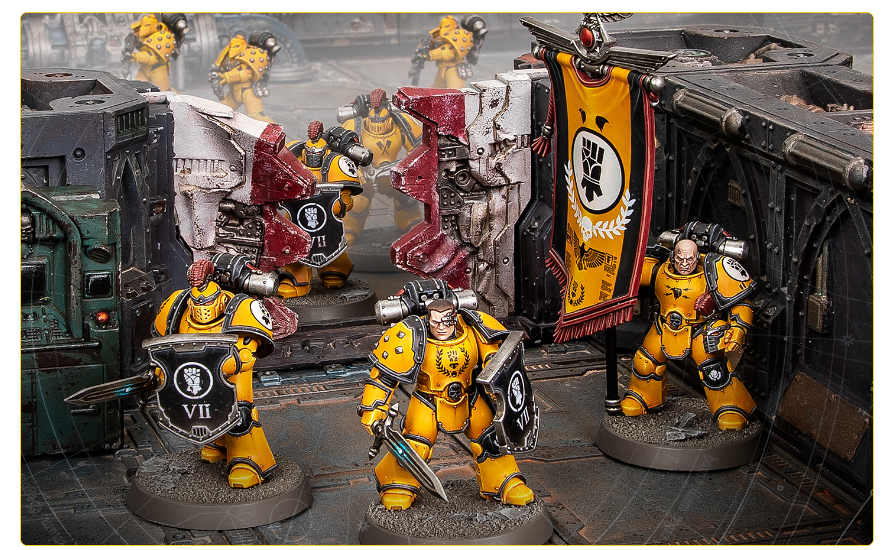 Yellow-armored Space Marines form a battle line on a sci-fi diorama with a black banner featuring a white fist emblem and ruined scenery behind them.