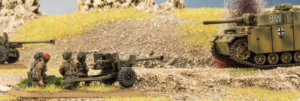 Soldiers load a towed field gun as a camouflaged tank rolls along a dirt road in the background.