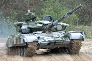 Green military tank driving on a dirt road through a forest, with the long main gun elevated and crew visible in the hatches thereon.