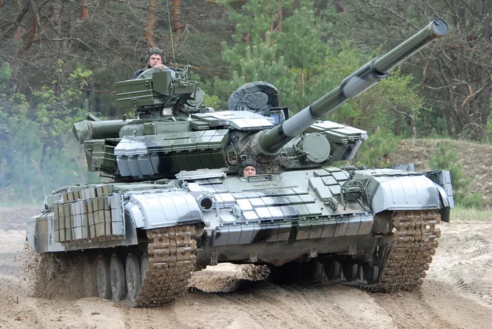 T64BV Frontline Gaming Green military tank driving on a dirt road through a forest, with the long main gun elevated and crew visible in the hatches thereon.