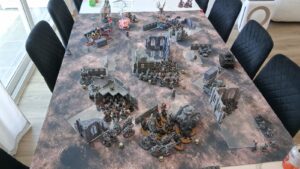 Tabletop wargame battle setup with multiple miniature armies and ruined city scenery on a large table.