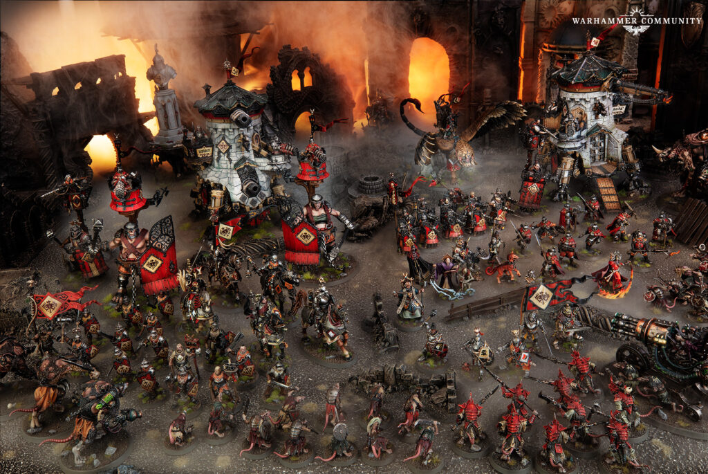 Massive Warhammer tabletop battle with red-armored squads, siege engines, and lava-lit ruins in the background; Warhammer Community logo visible.
