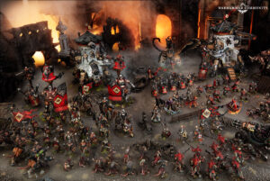 Massive Warhammer tabletop battle with red-armored squads, siege engines, and lava-lit ruins in the background; Warhammer Community logo visible.