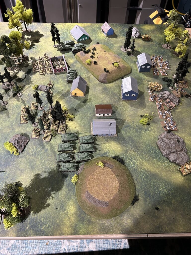 Tabletop wargame scene with a line of green tanks advancing toward a cluster of small houses and trees on a grassy battlefield.