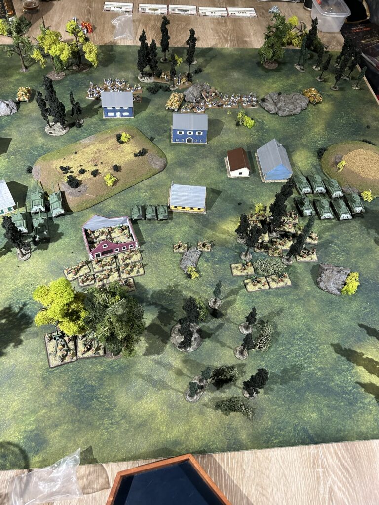 Tabletop war game scene with blue buildings, tanks, and infantry on a grassy battlefield with trees and hills.