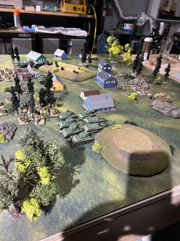 Tabletop wargame battlefield with green tanks advancing past blue houses, trees, and a large grassy hill in the foreground.