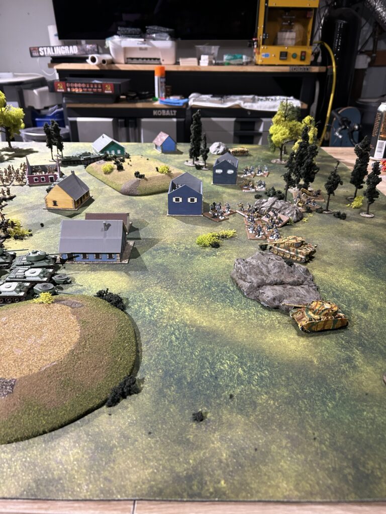 Tabletop wargame scene with miniature tanks, infantry, and small buildings on a grassy battlefield with trees and hills.
