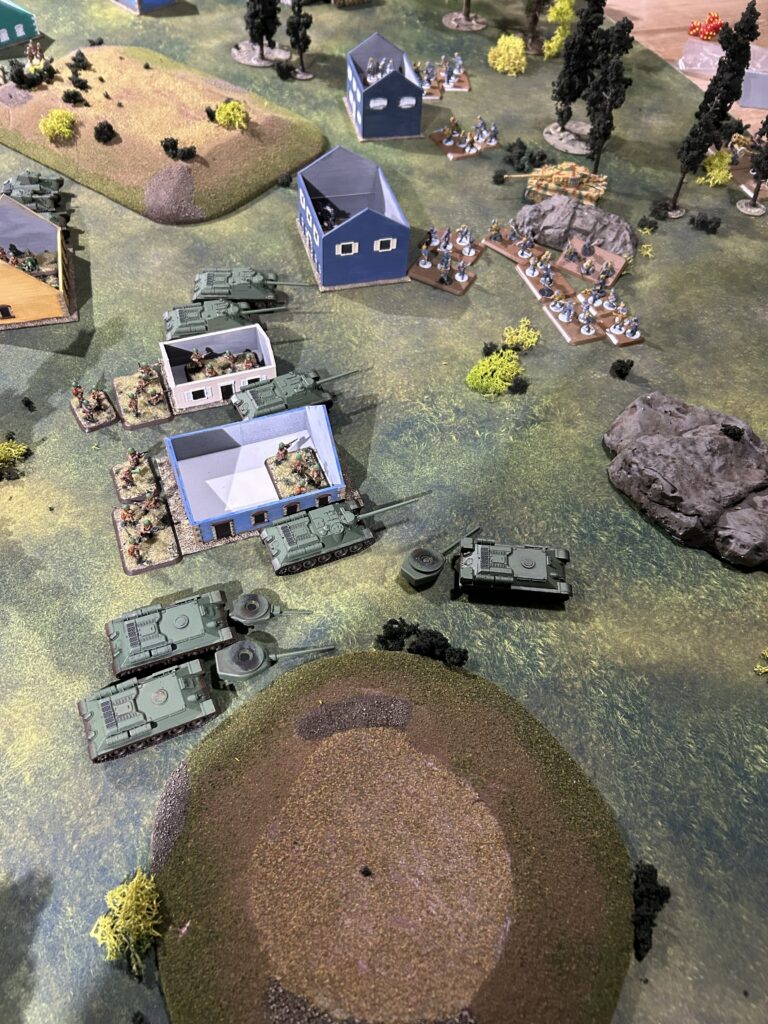 Tabletop war game scene showing green armored tanks advancing across a grassy battlefield, with blue rectangular buildings, groups of infantry, and scattered trees and hills.