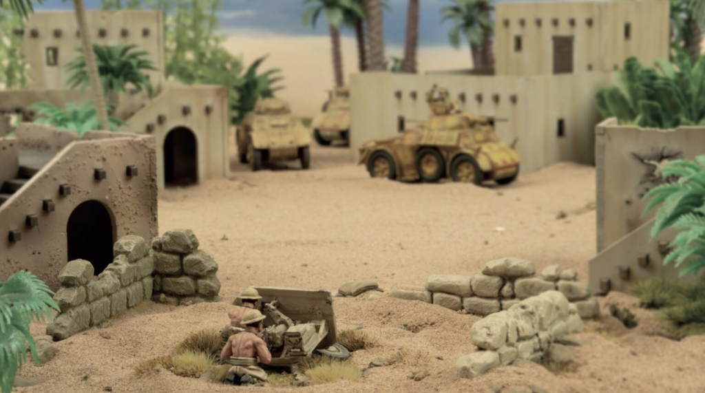 Miniature desert village scene with two soldiers operating artillery near a stone wall, tan buildings and armored vehicles in the background.
