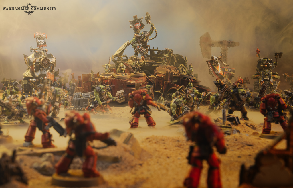 image Frontline Gaming Warhammer miniature battle scene with orc and human troops firing around a wrecked vehicle in a dusty battlefield with smoke and banners.