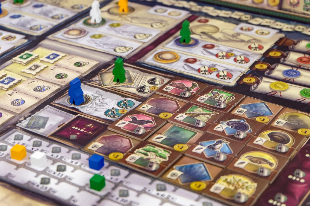 image Frontline Gaming A colorful board game setup with modular tile boards, cards, and multicolored plastic meeples (blue, green, yellow) placed across the grided play area.