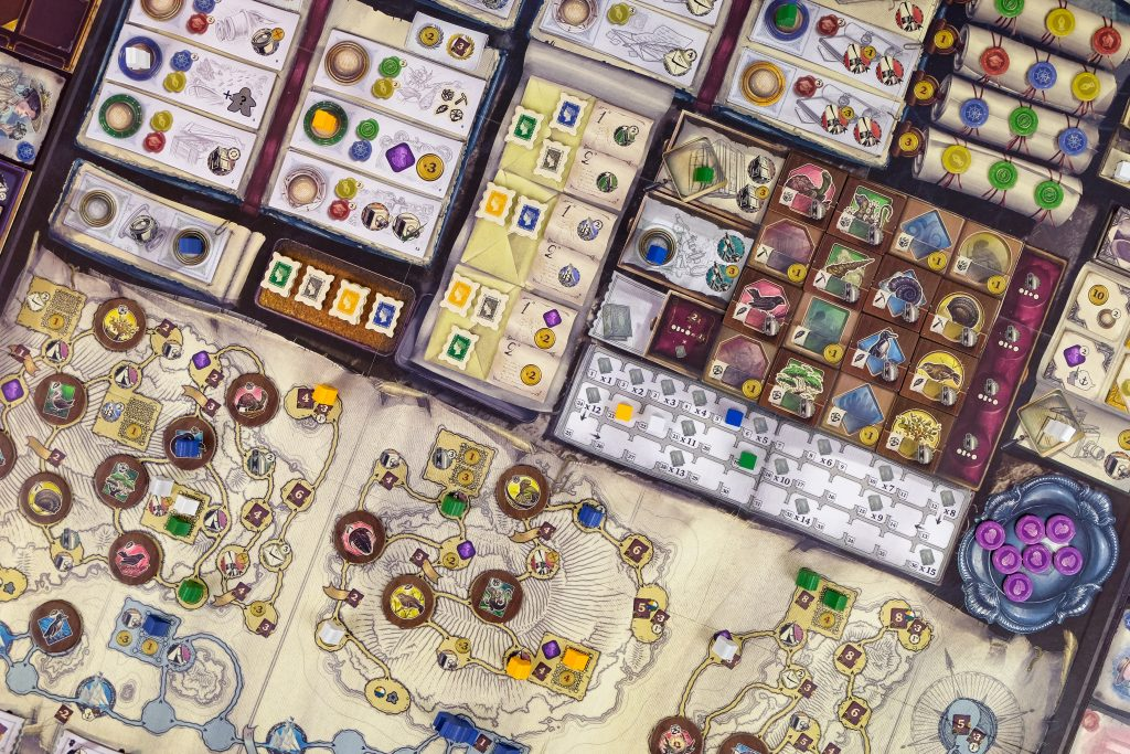 image Frontline Gaming Top-down view of a complex strategy board game with colorful tiles, tokens, and map-like panels spread across a tabletop.