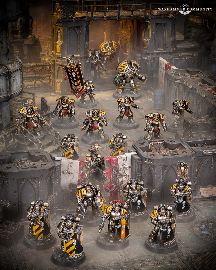 Tabletop Warhammer diorama: yellow-and-black armored miniatures advance through a rusted industrial battlefield with a banner at the center.