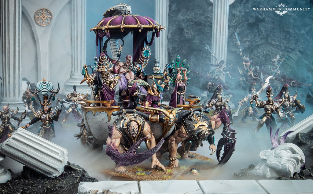 Warhammer miniature battle scene: a gilded chariot with ornate riders leads armored warriors through ruins, with blue fog and pillars in the background.
