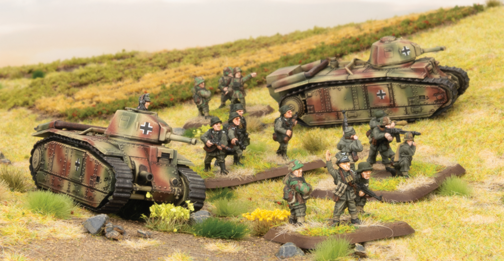 Scale model World War II battlefield: two camouflaged German tanks and a group of soldiers advancing across a grassy diorama.