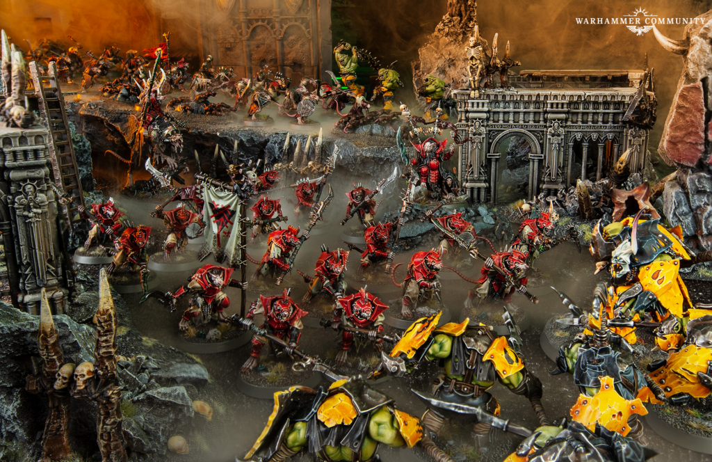 Warhammer miniature battle: red-armored Chaos troops clash with green orcs amid ruined Gothic ruins and smoky battlefield.