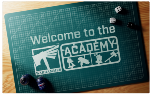 Green cutting mat showing Welcome to the Academy with the Warhammer logo and small hobby silhouettes in a game studio setup
