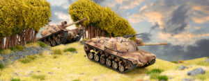 Two camouflaged toy tanks traverse a grassy rocky hill beside a row of trees under a cloudy sky model diorama