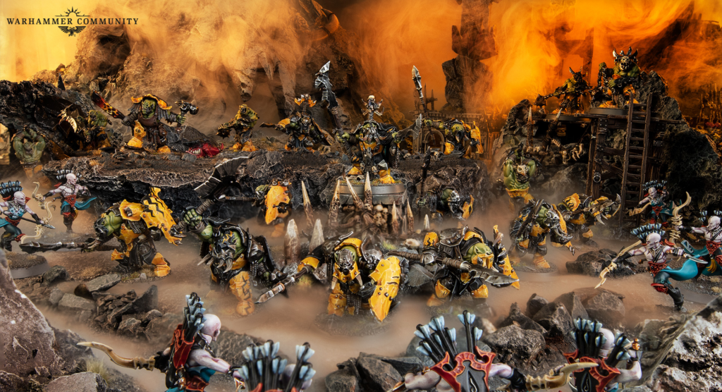 Warhammer battle scene with orcs and monsters clashing on a lava-lit rocky battlefield; Warhammer Community logo in the top left.