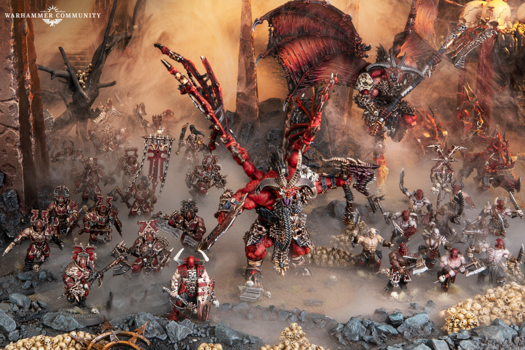 image Frontline Gaming Chaos Warhammer battle: red-armored Chaos Space Marines clash with a towering demon amid lava-lit ruins, skulls and jagged rocks in a smoky battlefield.