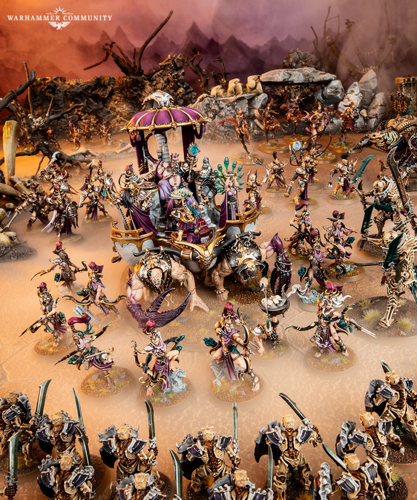 image Frontline Gaming Large Warhammer tabletop battle scene with dozens of intricately painted miniatures clashing on a dusty battlefield; a central ornate purple carriage is surrounded by armored figures and banners amid smoky rocks.