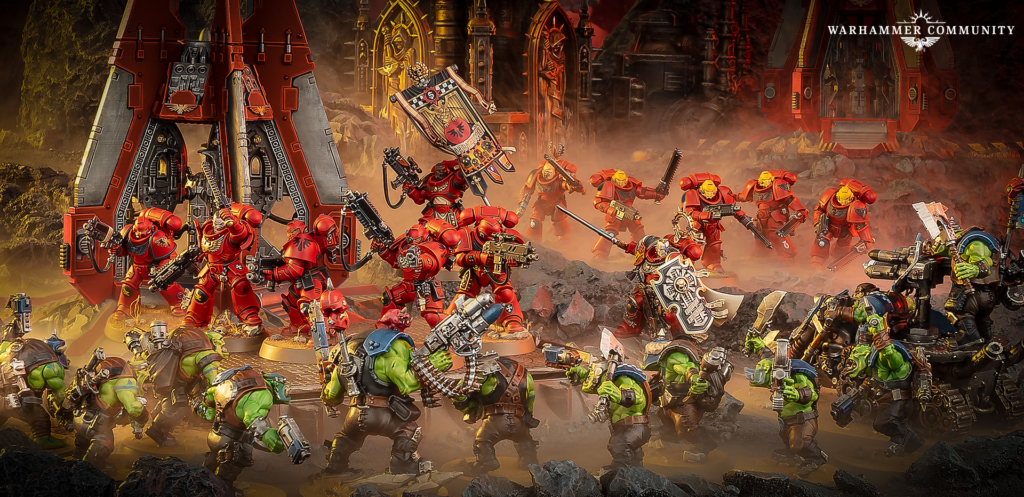 Red armored Space Marines fight green orc troops on a smoky rocky battlefield with towering red war machines in the background Warhammer Community logo visible in the top right