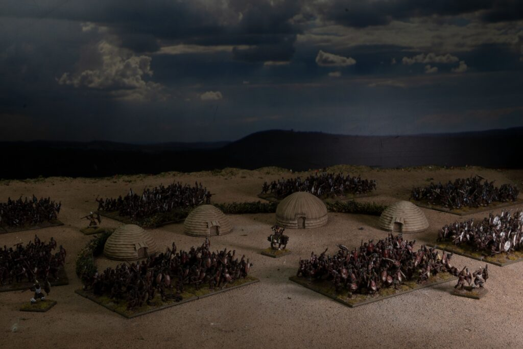 image Frontline Gaming Miniature battlefield with clustered dome huts and rows of tiny soldiers under a cloudy sky in a desert scene