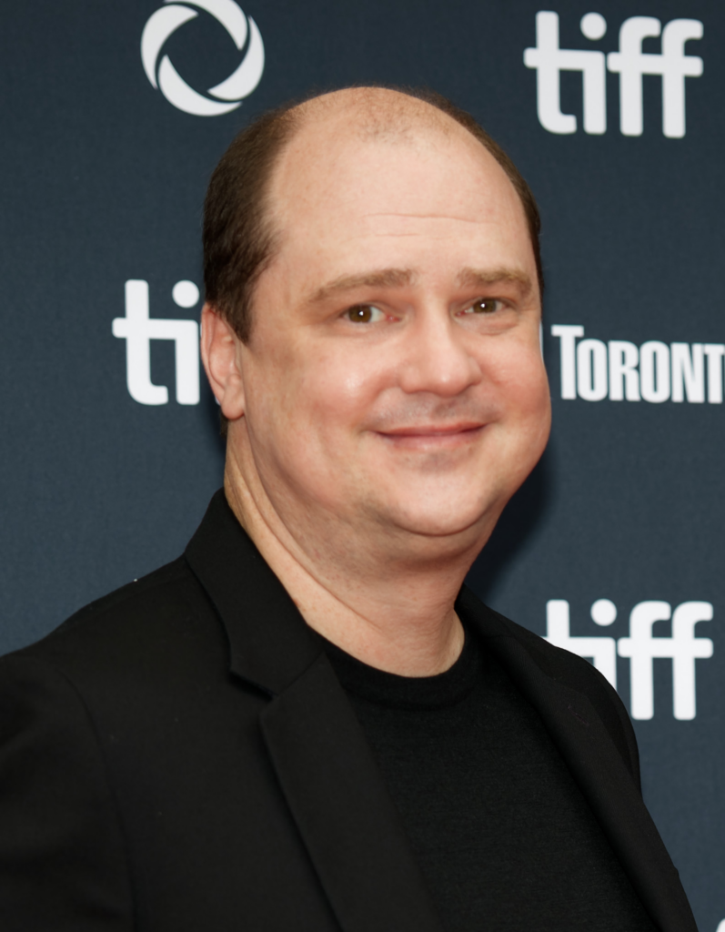 Smiling man posing for a photo at a TIFF event with a dark backdrop featuring white logos
