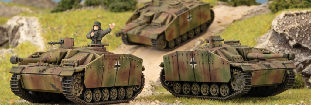 Two German camouflaged scale-model tanks face off on a dirt road in a rocky countryside diorama, with a soldier figure in one turret and a third tank in the background.