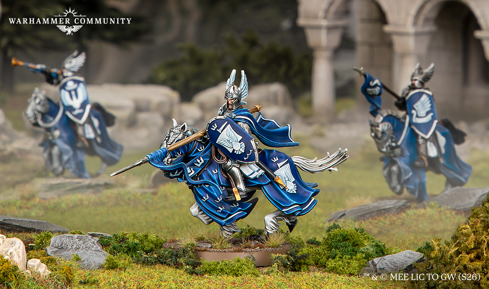 image Frontline Gaming Blue-armored Warhammer miniatures charging with spears and shields across a rocky, grassy battlefield, cloaks billowing; ruins in the background (Warhammer Community watermark in top left).