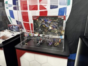 Display case with Gundam model kits and action figures on shelves including blue and red mecha boxes and scattered small figures in front of the boxes