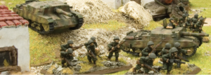 Miniature battlefield diorama showing green toy soldiers advancing toward two tanks amid rubble and grass