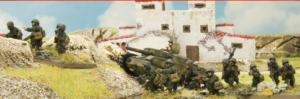 Scale model battlefield diorama with soldiers advancing and a tank near a white ruined building on a grassy terrain