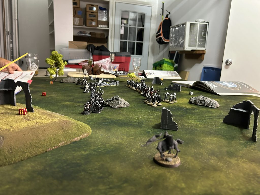 Tabletop wargaming scene with miniature troops advancing across a grassy battlefield dice on the table and cluttered background shelves and boxes in a room