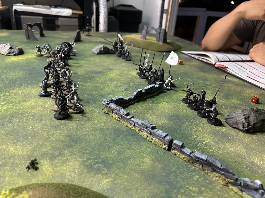 Tabletop wargaming scene with groups of miniature soldiers facing off on a grassy gaming mat ruins and walls scattered around a white die nearby and an open rulebook on the right