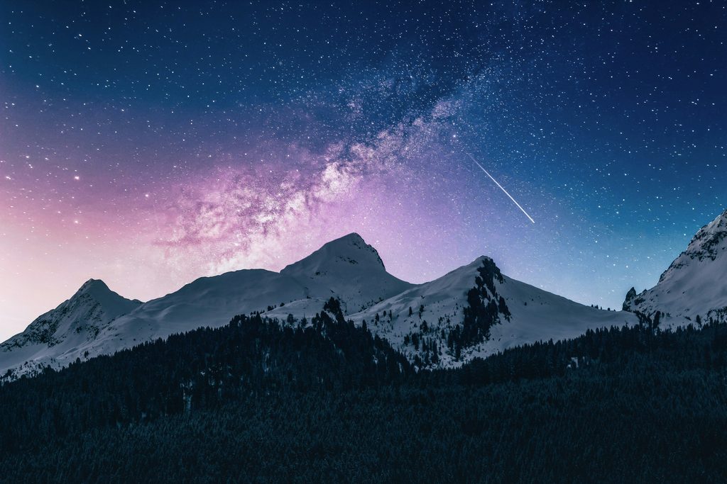 Snow covered mountain range at night under a star filled sky with the Milky Way and a shooting star streaking across