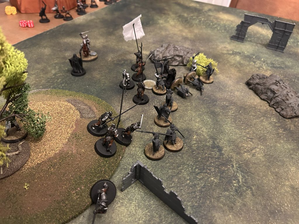 Tabletop wargame scene with armored miniatures engaging near a ruined wall and rocky terrain on a grassy battle mat