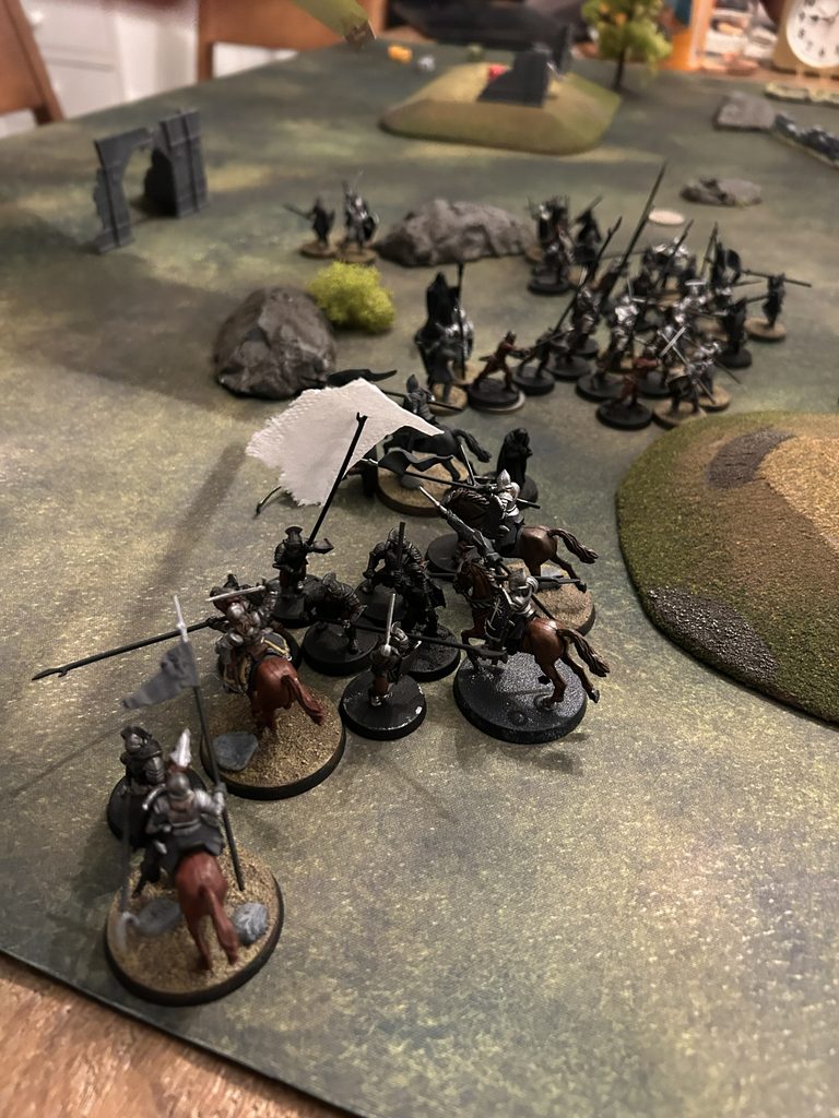 Tabletop wargame scene with mostly black armored infantry and cavalry advancing across a grassy battleboard with rocks a ruin arch bushes and rolling hills in the background