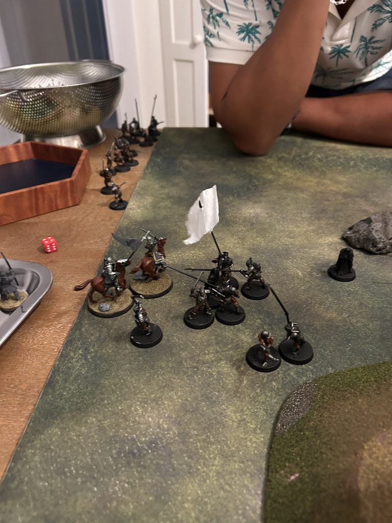 Miniature soldiers in a skirmish on a textured tabletop battlefield with a red die nearby and terrain pieces in view