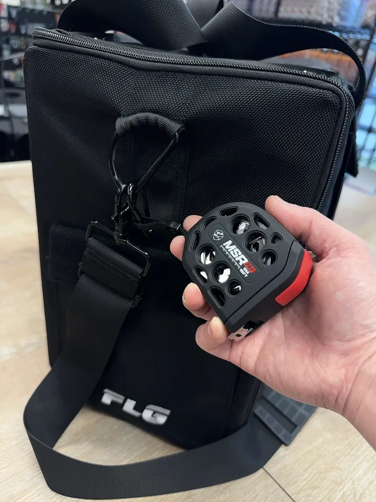 flgtapemeasure1 Frontline Gaming Close up of a hand holding a small black luggage lock with round dials attached to a black bag zipper